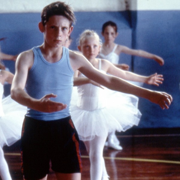 Billy Elliot tanzt mit grimmigem Gesichtsausdruck inmitten einer Gruppe Ballettänzerinnen.