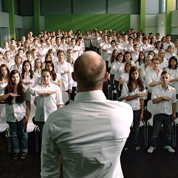 Ein Lehrer steht vor seiner Klasse. Alle Schüler*innen tragen Uniform und führen denselben Gruß aus.
