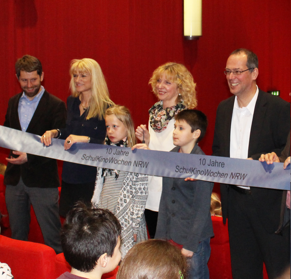 Die Organisatoren und Teilnehmer*innen stehen in einem Kinosaal und halten ein langes Banner mit der Aufschrift "10 Jahre SchulKinoWochen NRW".
