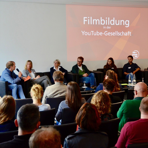 Ein Podium aus sechs Sprechern und vier Reihen von Zuschauer:innen sitzen vor einer Leinwand mit dem Text "Filmbildung in der YouTube-Gesellschaft".