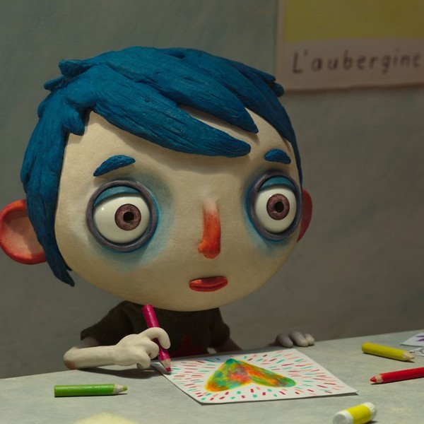 Ein Junge aus Knete mit blauen Haaren sitzt an einem Tisch und malt ein Bild.