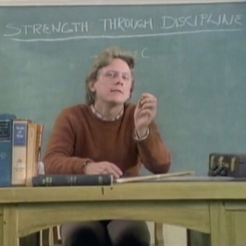Ein strenger Lehrer sitzt am Pult vor einer Tafel auf der steht "strength through discipline".