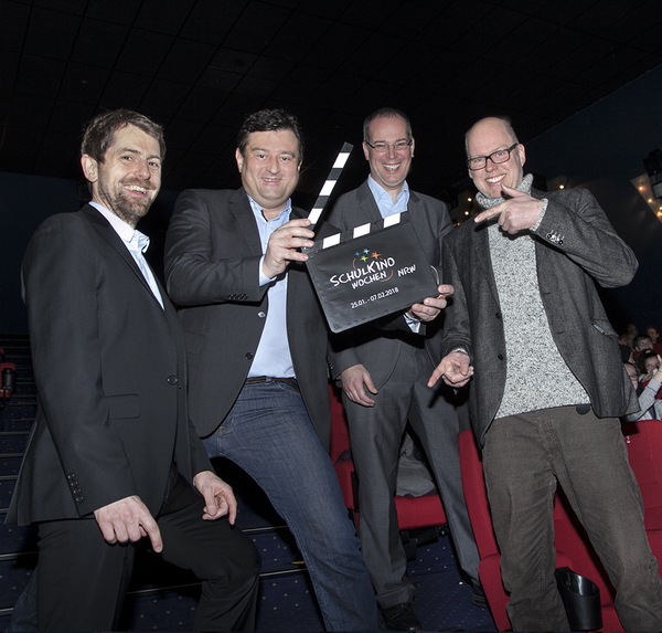 Vier Männer stehen in einem Kinosaal, einer hält eine Filmklappe mit der Aufschrift "SchulKinoWochen NRW"