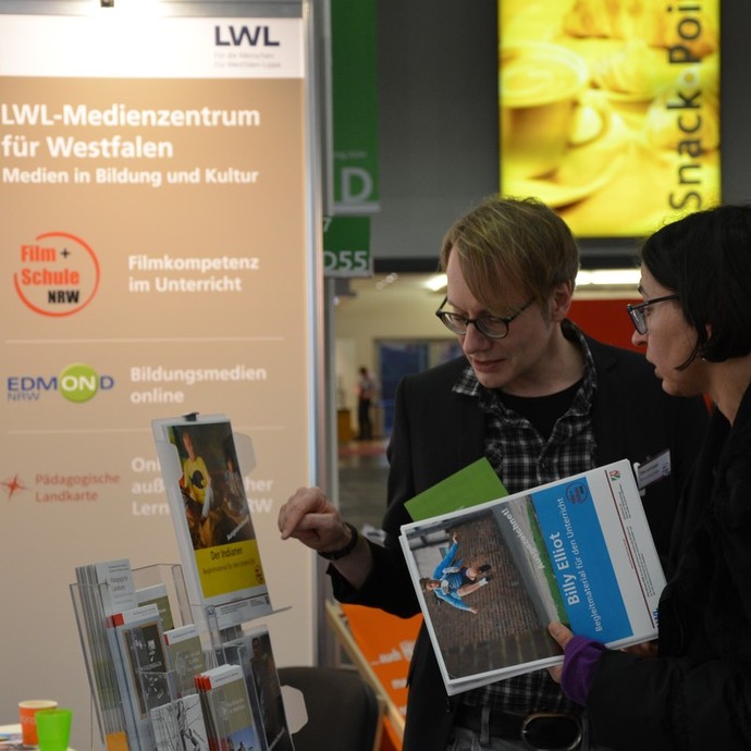 Menschen schauen sich das Material von FILM+SCHULE an ihrem Stand an. (vergrößerte Bildansicht wird geöffnet)