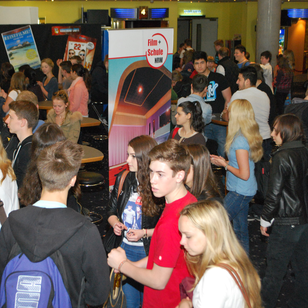 Eine Gruppe Schüler*innen in einem Kino Foyer, in der Mitte ein Aufsteller von FILM+SCHULE NRW.