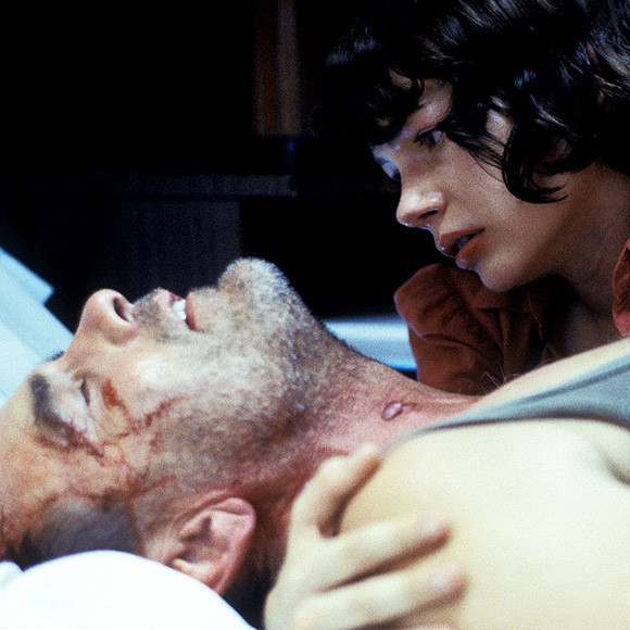 Eine junge Frau hält einen Mann im Arm, der unter Schmerzen auf dem Bett liegt.