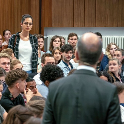Eine Studentin und ein Professor stehen in einem Hörsaal voller Studierender.