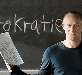 Ein Lehrer steht vor einer Tafel auf der das Wort "Autokratie" steht.