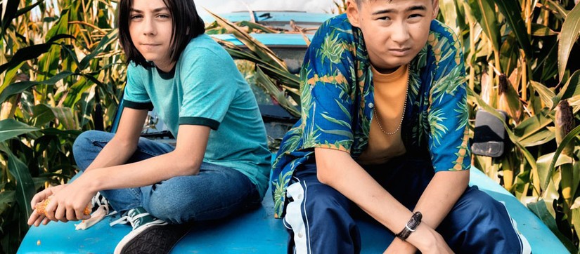 Zwei Jungen sitzen auf der Motorhaube eines Autos mitten in einem Maisfeld und gucken in die Kamera.