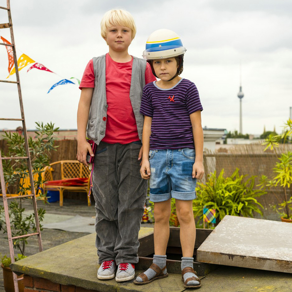 Ein Junge mit blonden Haaren und ein kleinerer Junge mit einem Fahrradhelm stehen nebeneinander auf einer Dachterasse.