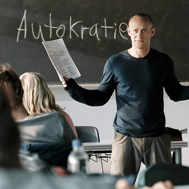 Filmstill aus Die Welle: Ein Lehrer steht vor einer Klasse. Hinter ihm auf der Tafel steht das Wort "Autokratie".