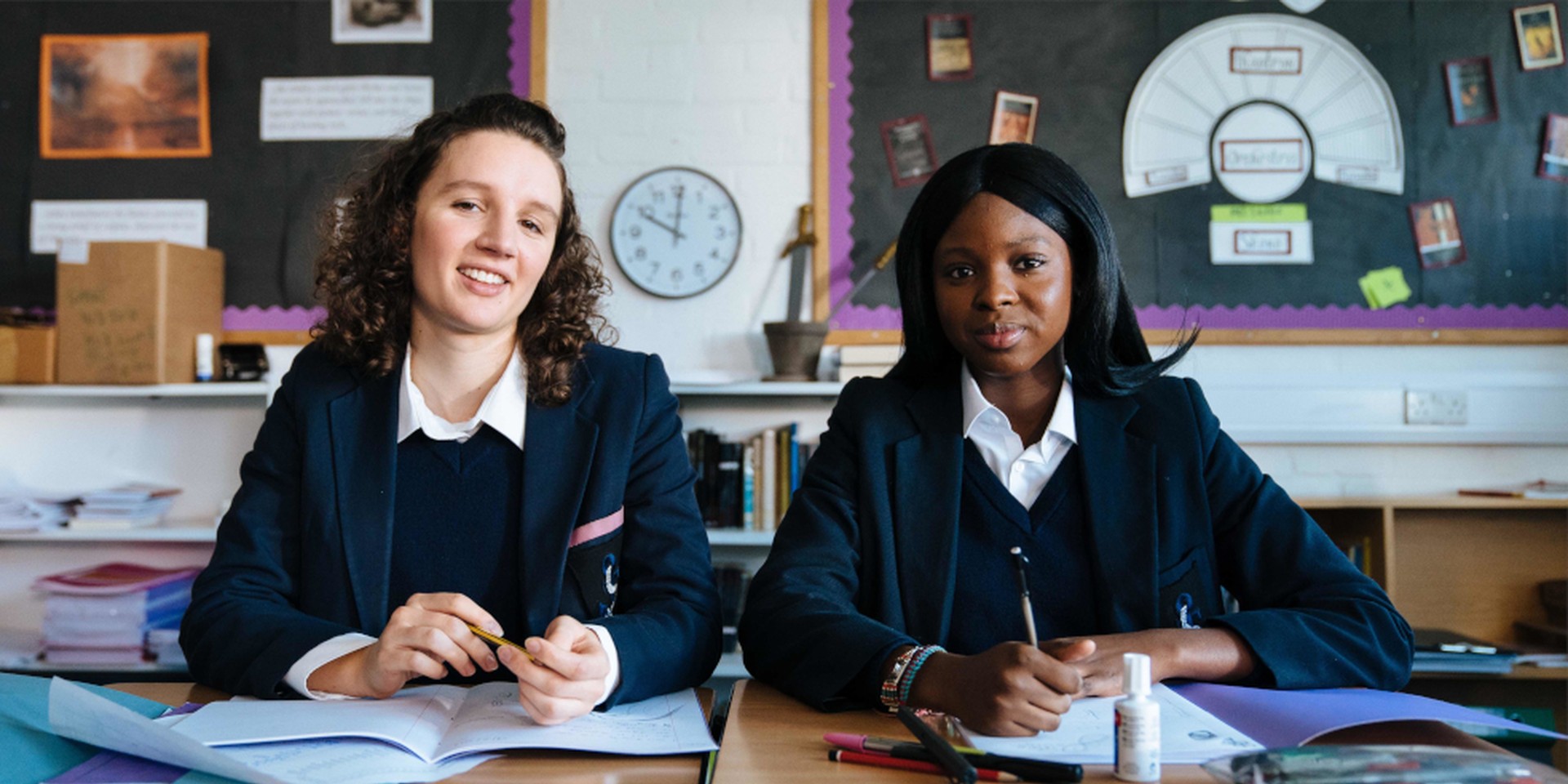 Zwei Mädchen mit englischen Schuluniformen sitzen in einer Schulklasse.