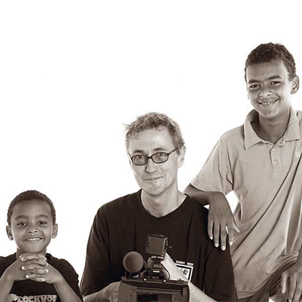 Ein Mann mit einer Kamera und zwei Jungen vor einem weißen Hintergrund.