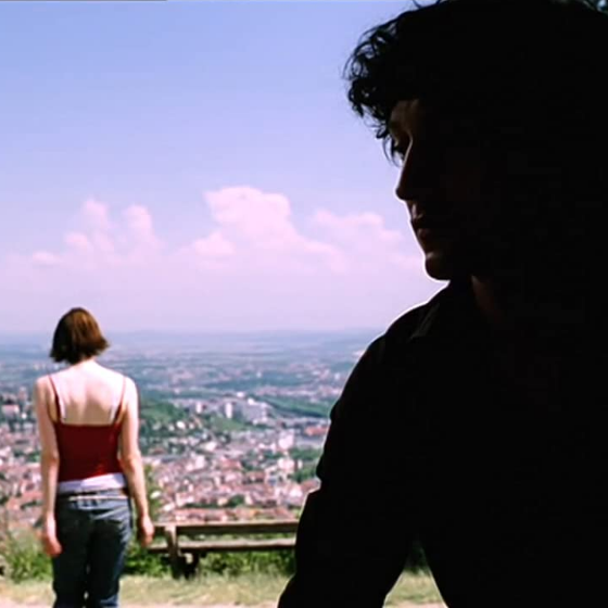 Ein Mann sitzt im Schatten. Hinter ihm läuft eine Frau in der Sonne einen Berg hinunter in Richtung einer Stadt.