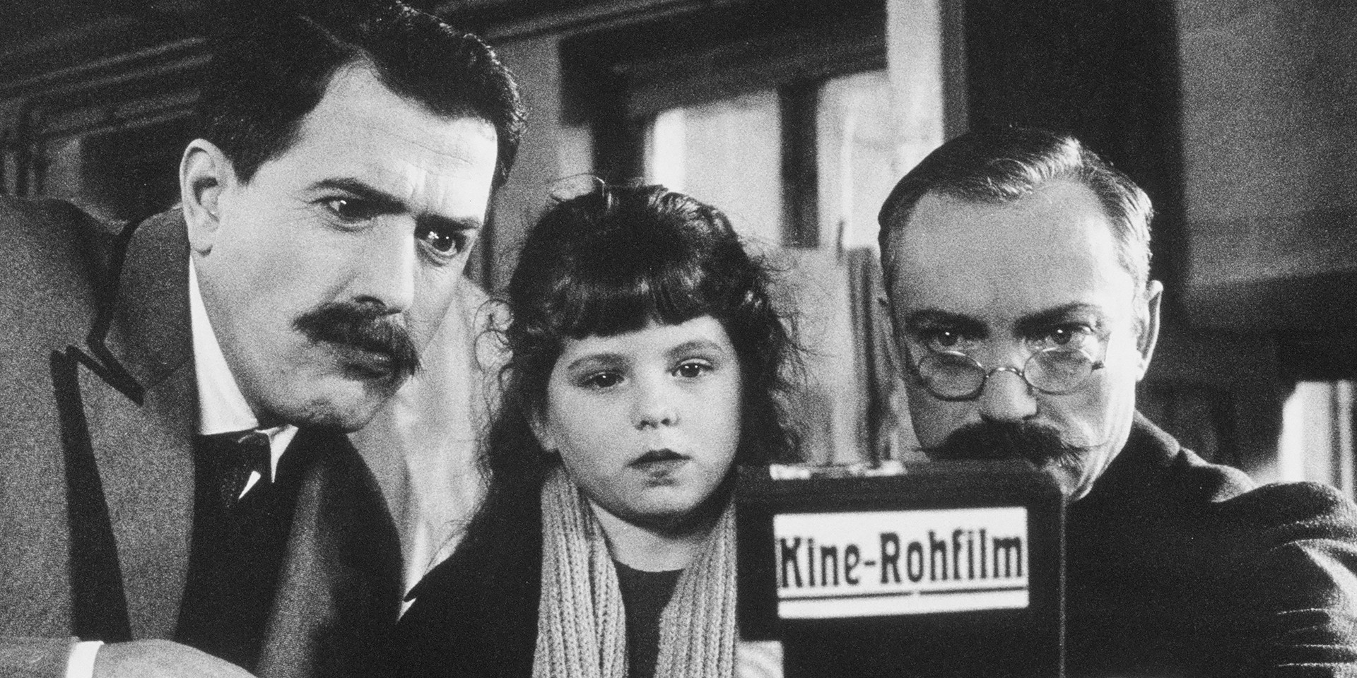 Zwei Männer und ein Mädchen schauen auf eine Schachtel mit der Aufschrift "Kine-Rohfilm".